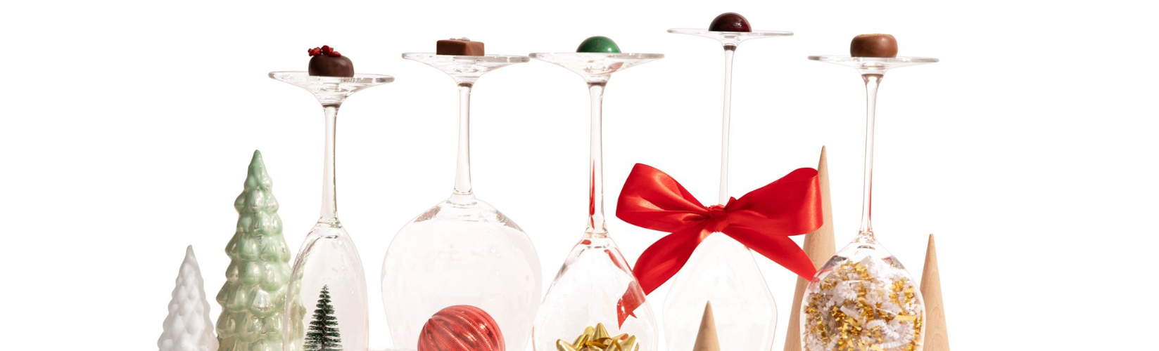 Upside-down wine glasses with assorted chocolates on top, surrounded by festive decorations including small Christmas trees and cones. One glass has a red bow tied around the stem.